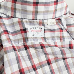 May include: Close-up of a plaid shirt in white, red, black, and gray. The shirt has a collar and a label that reads "ARROW CONTOUR TAILORED WASH AND WEAR 100% COTTON". The shirt is made in the U.S.A.