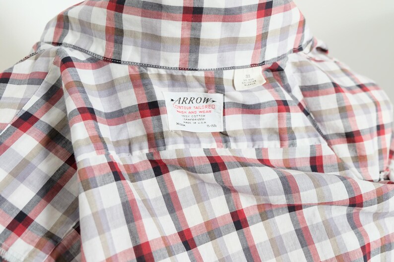 May include: Close-up of a plaid shirt in white, red, black, and gray. The shirt has a collar and a label that reads "ARROW CONTOUR TAILORED WASH AND WEAR 100% COTTON". The shirt is made in the U.S.A.
