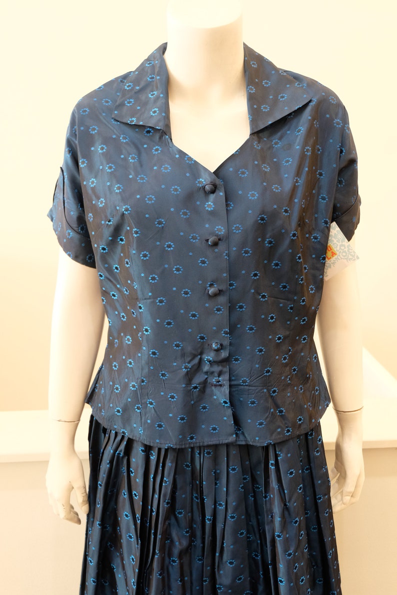 K&ouml;nnte beinhalten: Zweiteiliges marineblaues Outfit auf einer Schaufensterpuppe. Das Oberteil ist eine Knopfbluse mit kurzen &Auml;rmeln und Kragen, verziert mit einem sich wiederholenden blauen Blumenmuster. Der passende Rock ist plissiert und hat das gleiche Blumenmuster.