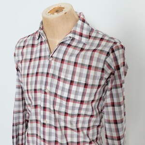 May include: A long-sleeved plaid shirt in a red, black, and white checkered pattern. The shirt is buttoned and has a chest pocket. The shirt is displayed on a mannequin.