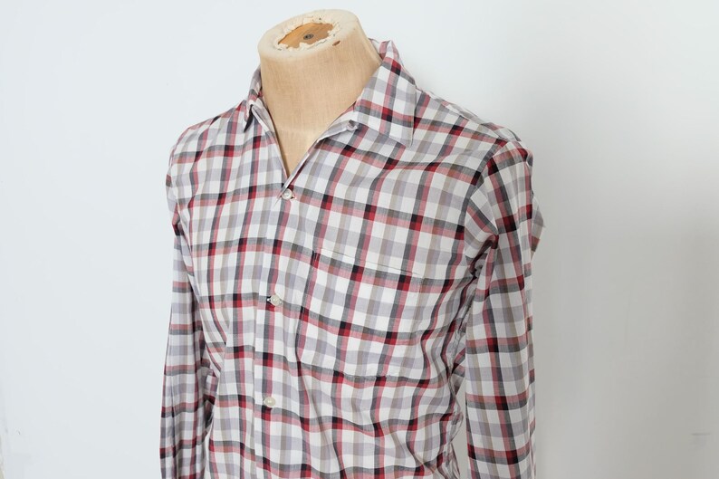 May include: A long-sleeved plaid shirt in a red, black, and white checkered pattern. The shirt is buttoned and has a chest pocket. The shirt is displayed on a mannequin.