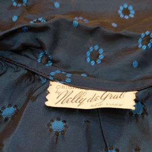 K&ouml;nnte beinhalten: Nahaufnahme eines dunkelblauen Stoffes mit einem Blumenmuster in Blau und Schwarz. Der Stoff ist am Halsausschnitt gerafft, und ein Etikett lautet "ORIGINAL by Nelly de Grab NEW YORK."