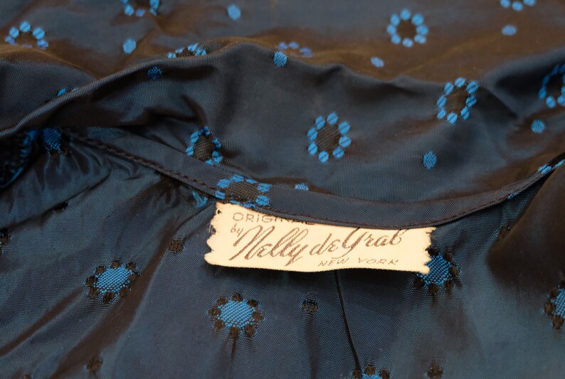 K&ouml;nnte beinhalten: Nahaufnahme eines dunkelblauen Stoffes mit einem Blumenmuster in Blau und Schwarz. Der Stoff ist am Halsausschnitt gerafft, und ein Etikett lautet "ORIGINAL by Nelly de Grab NEW YORK."