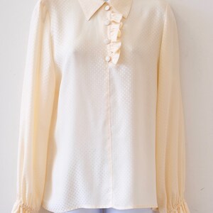 May include: A cream-colored, long-sleeved blouse with a ruffled neckline and a button-up front. The blouse has a subtle polka dot pattern.