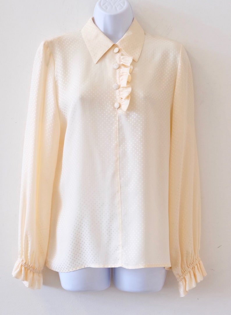 May include: A cream-colored, long-sleeved blouse with a ruffled neckline and a button-up front. The blouse has a subtle polka dot pattern.
