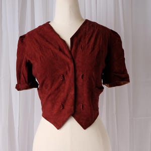 K&ouml;nnte beinhalten: Burgunderfarbene Kurzarmjacke mit V-Ausschnitt und doppelreihigem Knopfverschluss. Die Jacke hat eine taillierte Passform und wird auf einer Schaufensterpuppe pr&auml;sentiert. Die &Auml;rmel sind umgeschlagen, und der Stoff hat eine strukturierte Oberfl&auml;che.