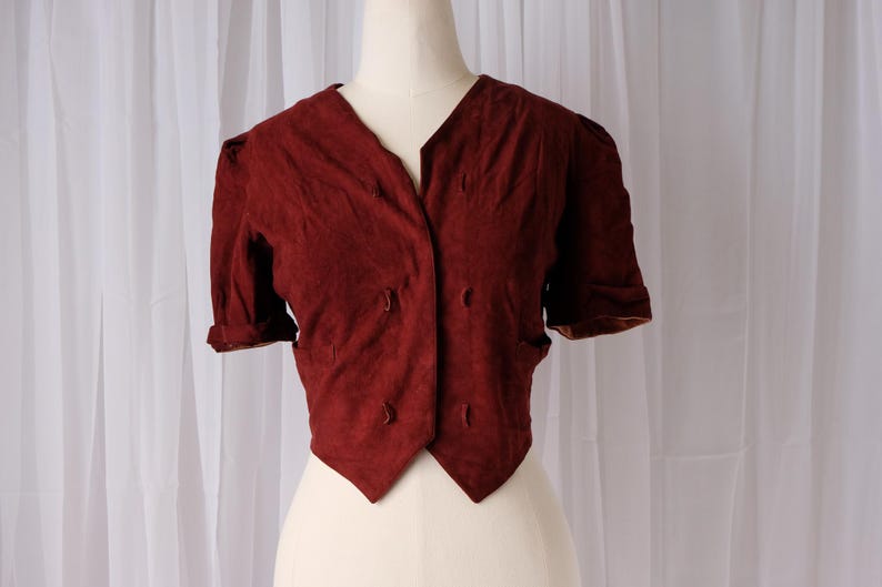 K&ouml;nnte beinhalten: Burgunderfarbene Kurzarmjacke mit V-Ausschnitt und doppelreihigem Knopfverschluss. Die Jacke hat eine taillierte Passform und wird auf einer Schaufensterpuppe pr&auml;sentiert. Die &Auml;rmel sind umgeschlagen, und der Stoff hat eine strukturierte Oberfl&auml;che.