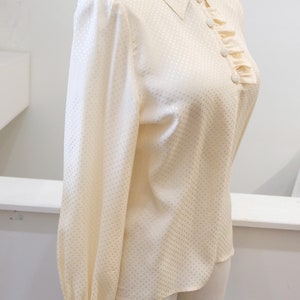 May include: A cream-colored, long-sleeved blouse with a white polka dot pattern. The blouse has a ruffled collar and a button-up front.