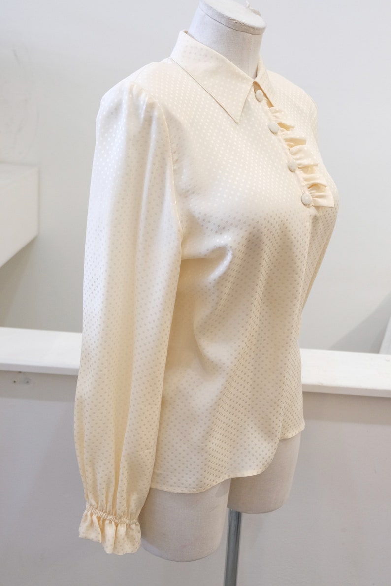 May include: A cream-colored, long-sleeved blouse with a white polka dot pattern. The blouse has a ruffled collar and a button-up front.