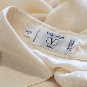 May include: A white silk blouse with a small white polka dot pattern. The blouse has a collar and a label that reads "valentino" and "Made in Italy".