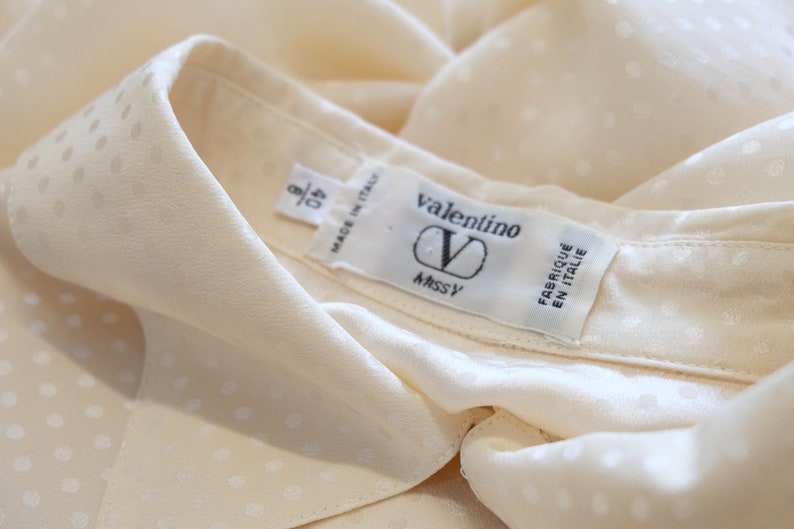 May include: A white silk blouse with a small white polka dot pattern. The blouse has a collar and a label that reads "valentino" and "Made in Italy".