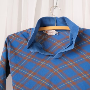 May include: A blue long-sleeved shirt with a plaid pattern in brown and yellow. The shirt has a collared neckline and is hanging on a wooden hanger. A tag is visible near the collar.