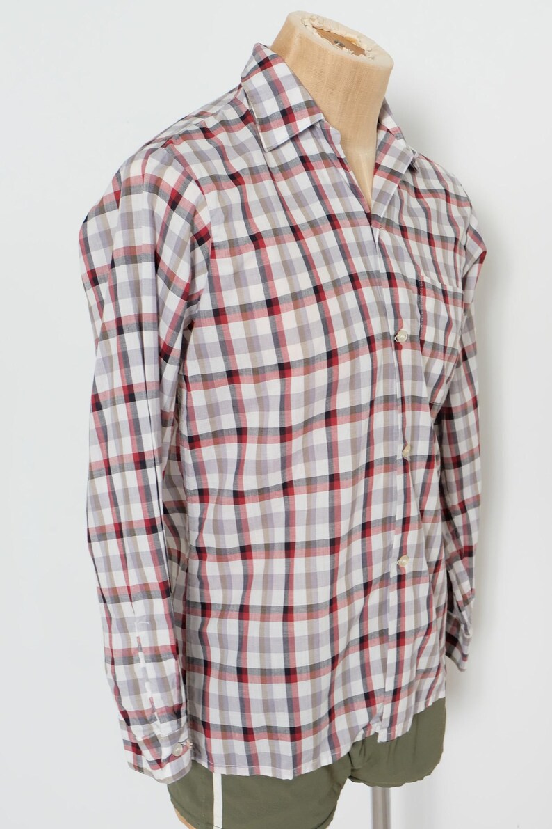 May include: A long-sleeved button-down shirt with a plaid pattern in white, gray, red, and black. The shirt is displayed on a mannequin and has a classic collar and buttoned cuffs. The shirt is paired with olive green shorts.