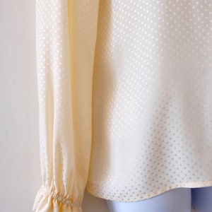 May include: A cream-colored blouse with a white polka dot pattern. The blouse has long sleeves with ruched cuffs.