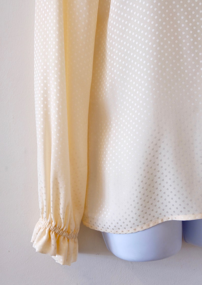 May include: A cream-colored blouse with a white polka dot pattern. The blouse has long sleeves with ruched cuffs.