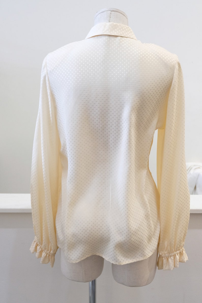 May include: A white long-sleeved blouse with a small white polka dot pattern. The blouse has a ruffled collar and cuffs.