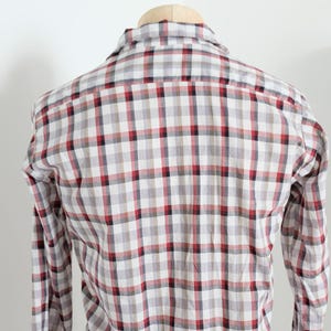 May include: A long-sleeved plaid shirt in a neutral color palette of white, gray, and red. The shirt is displayed on a wooden hanger against a plain white background. The plaid pattern is a classic design.
