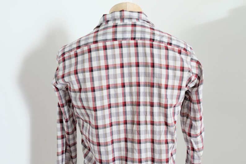 May include: A long-sleeved plaid shirt in a neutral color palette of white, gray, and red. The shirt is displayed on a wooden hanger against a plain white background. The plaid pattern is a classic design.