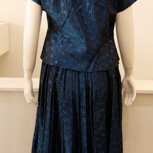 K&ouml;nnte beinhalten: Marineblaues zweiteiliges Outfit mit einem kurz&auml;rmeligen Oberteil und einem plissierten Midirock. Der Stoff ist mit kleinen, hellblauen Blumenmustern versehen. Das Outfit wird auf einer Schaufensterpuppe pr&auml;sentiert.