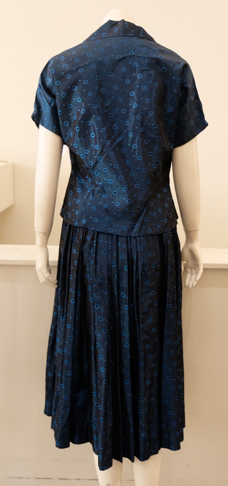K&ouml;nnte beinhalten: Marineblaues zweiteiliges Outfit mit einem kurz&auml;rmeligen Oberteil und einem plissierten Midirock. Der Stoff ist mit kleinen, hellblauen Blumenmustern versehen. Das Outfit wird auf einer Schaufensterpuppe pr&auml;sentiert.