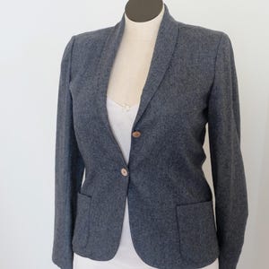 May include: A grey blazer with a shawl collar and two front pockets. The blazer has two wooden buttons and is displayed on a mannequin. A white top is visible underneath the blazer.