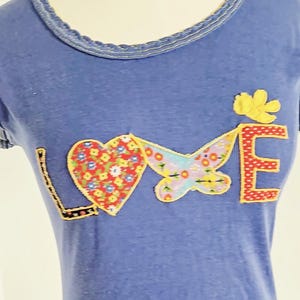May include: Blue t-shirt featuring the word "LOVE" appliquéd on the front. The "L" is black, the "O" is a heart with a floral pattern, the "V" is a butterfly, and the "E" is red polka dots. A yellow flower is above the "E".