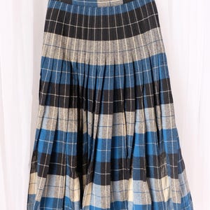 May include: A vintage pleated skirt in a blue, black, gray, and cream plaid pattern. The skirt hangs from a wooden hanger against a white backdrop. The skirt's design features a classic plaid pattern.