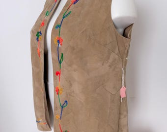May include: A tan suede vest with colorful floral embroidery along the edges. The embroidery features red, yellow, blue, and green thread. The vest is displayed on a white mannequin.