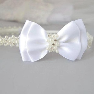 May include: A white satin bow headband with pearl and rhinestone detailing. The headband features a large, layered bow in the centre, adorned with small pearls and sparkling rhinestones. The band itself is covered in small, white pearls.
