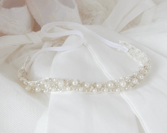 May include: A white pearl headband with a delicate ribbon tie. The headband is on a white fabric background.