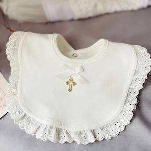 May include: White cotton bib with a lace trim and a gold cross charm. The bib has a white satin bow.