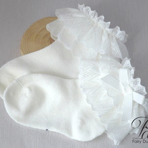May include: White cotton socks with a white lace ruffle and a white satin bow. The socks are on a wooden surface.
