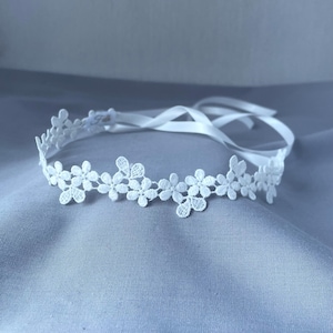 May include: A white lace floral headband with a satin ribbon tie. The headband features a repeating pattern of small, detailed flowers. The ribbon is white and the headband is approximately 2.5 cm wide.