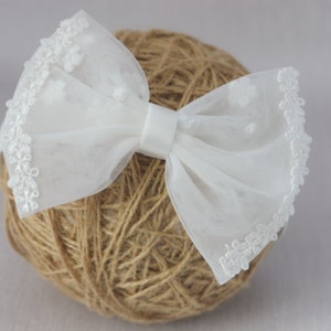 May include: A white tulle bow with a white lace trim. The bow is tied with a white satin ribbon and is sitting on a ball of twine.