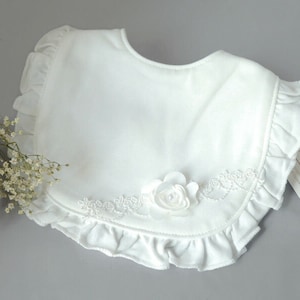 May include: White cotton baby bib with a ruffled edge and a white satin flower applique. The bib has delicate embroidery around the edge.