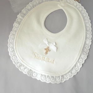May include: Ivory bib with a white lace trim and a small bow. A gold cross charm hangs from the bow. The name "Sienna" is embroidered in gold thread. A baby item.