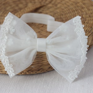 May include: A white tulle bow with a satin center and lace trim. The bow is attached to an elastic headband.