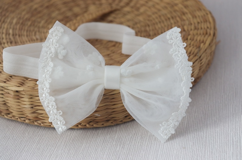 May include: A white tulle bow with a satin center and lace trim. The bow is attached to an elastic headband.