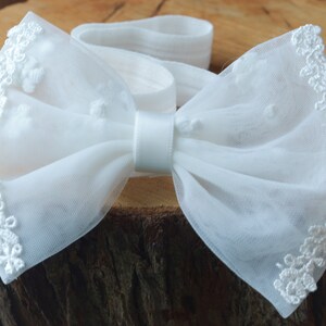 May include: White tulle bow with a satin center and lace trim. The bow is tied around a white elastic headband.