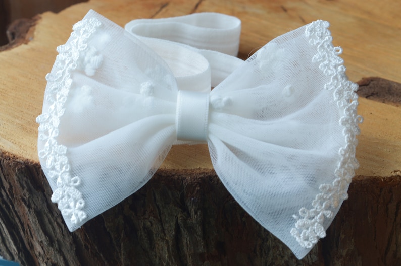 May include: White tulle bow with a satin center and lace trim. The bow is tied around a white elastic headband.
