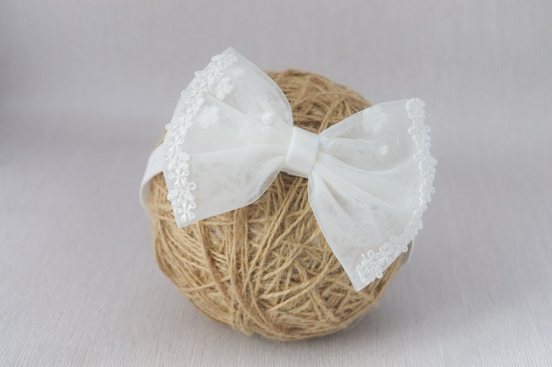 May include: White tulle bow headband with a delicate lace trim. The bow is tied in a simple knot and the headband is made of a soft elastic material.