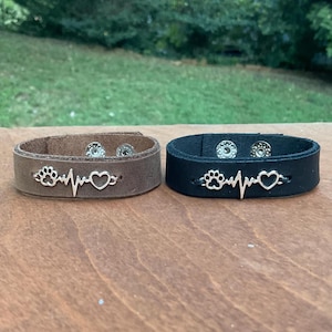 May include: Two leather bracelets with snap closures. One is brown and the other is black. Both feature a silver charm with a paw print, a heart, and a heartbeat line.