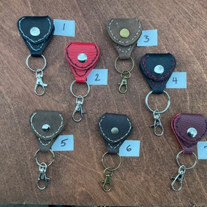 May include: Seven leather guitar pick holders with keychains. The holders are in various colors, including black, brown, red, and burgundy. Each holder has a small metal button on the front. The keychains are silver and have a spring clip.