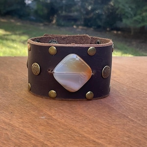 May include: A brown leather cuff bracelet with a large, white and orange agate stone in the center. The bracelet is decorated with brass studs and rivets.