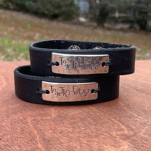 May include: Two black leather bracelets stacked on top of each other. Each bracelet has a silver plate with the words "Together Forever" engraved on it. The bracelets are on a wooden surface.