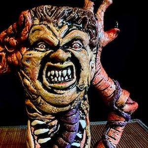 May include: A ceramic mug shaped like a monster with a grotesque face and a twisted body. The mug is painted in shades of brown, orange, yellow, and purple.