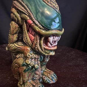 May include: A ceramic tiki mug shaped like a xenomorph creature with a smooth, elongated green head, exposed teeth, and intricate biomechanical textures in shades of brown, tan, and dark green. The figure stands on two feet with clawed toes.