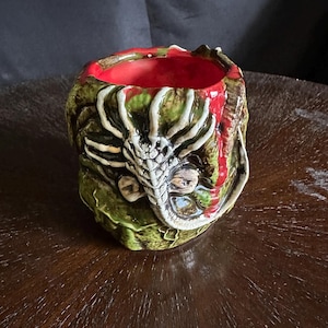 May include: A ceramic, hand-painted planter with a dragon design. The planter features a red interior, with a green and brown exterior. The dragon's head is white and gray, with a detailed, textured appearance. The planter is sitting on a dark wooden surface.
