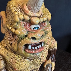 May include: A sculpted figurine of a cyclopean creature with yellow, textured, reptilian-like skin, a single central eye with a blue iris, a prominent horn on its forehead, and an open mouth revealing white teeth, accented with brown fur-like details.