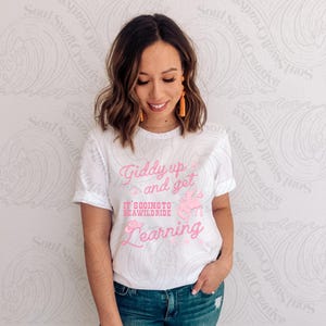 May include: White t-shirt with the text "Giddy up and get, it's going to be a wild ride, learning" in pink script. The shirt features a pink graphic of a horse and a cowboy hat. The model is wearing blue jeans.